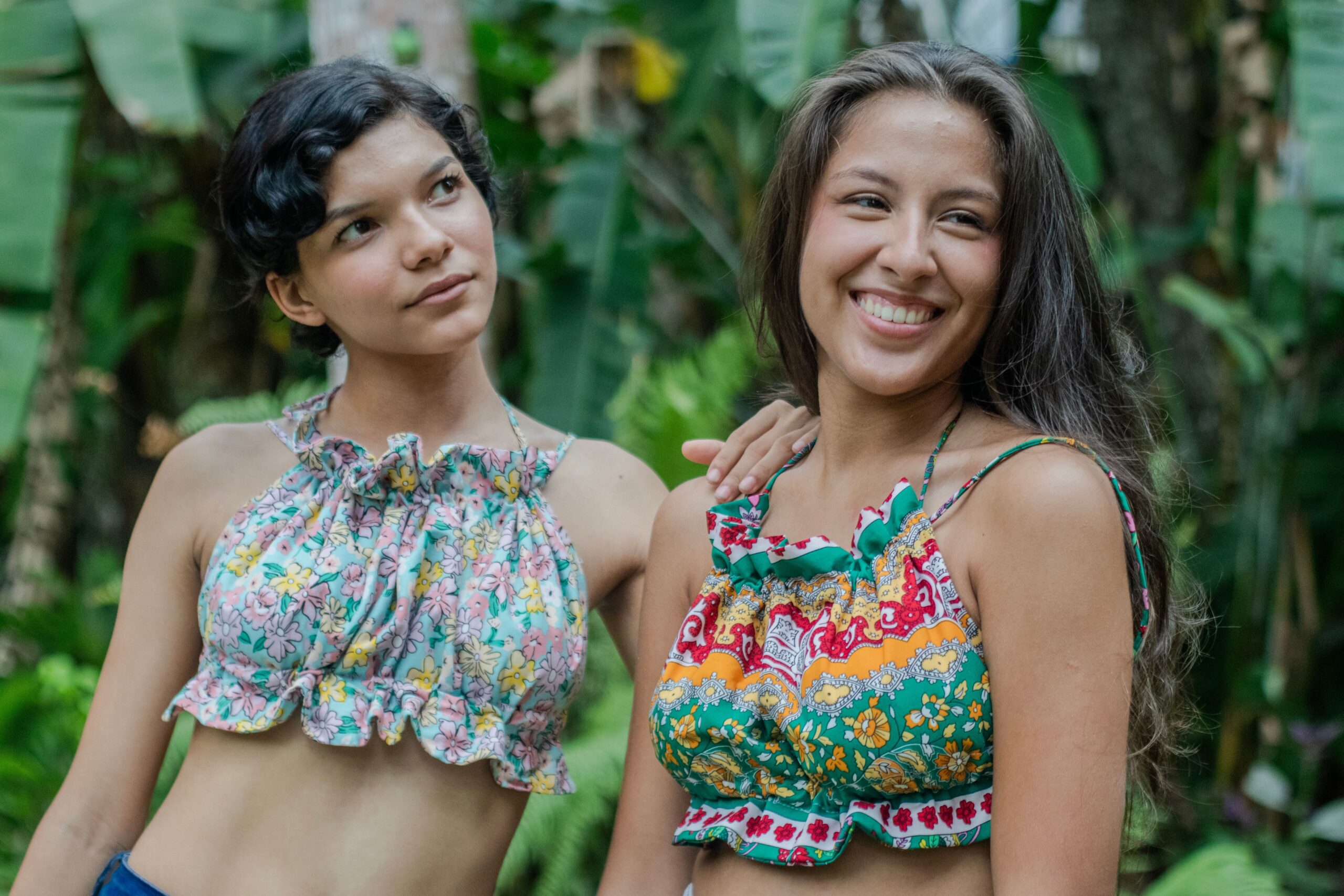 Two women in colorful summer tops enjoy outdoor fashion photography amidst lush greenery.